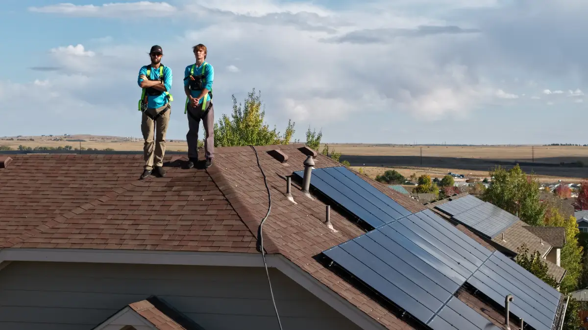 Two workers standing on a brown shingled roof with solar panels installed.