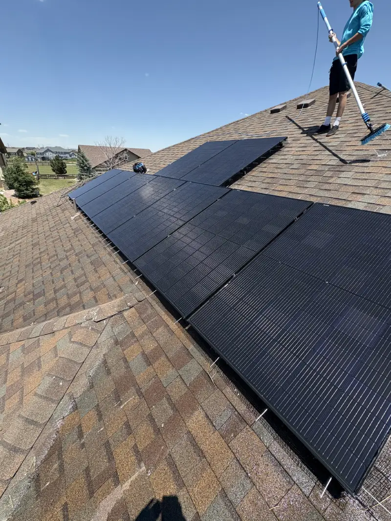 Person standing on a brown shingled roof cleaning large black solar panels with a long-handled brush under a clear blue sky.