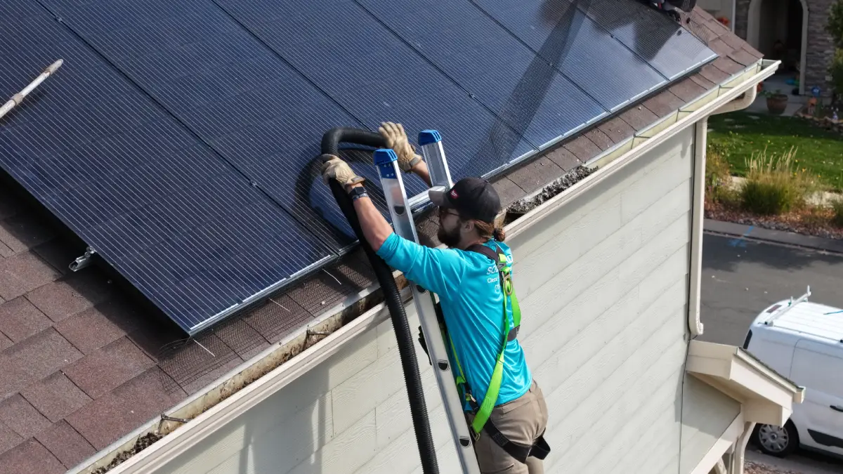 Worker wearing safety harness vacuuming debris from gutters below solar panels on a house roof.