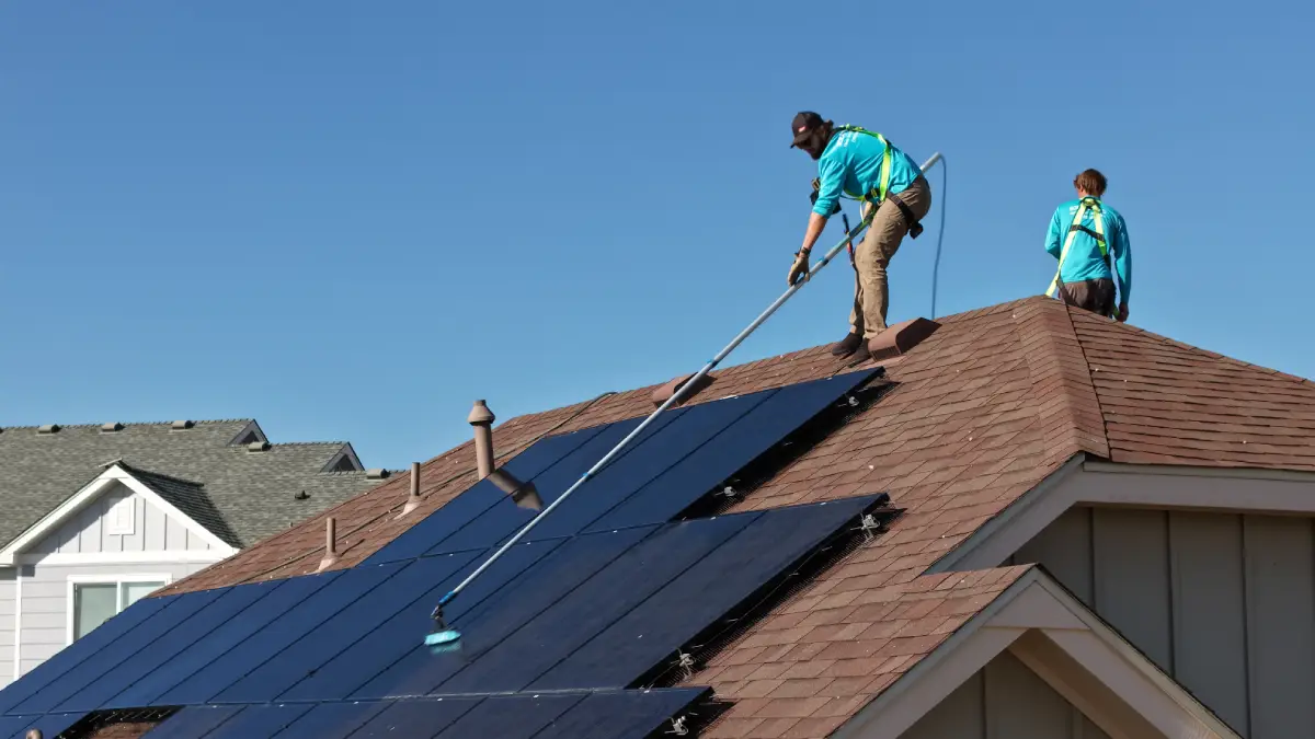 Two workers in blue shirts cleaning solar panels on a brown shingled roof under clear blue sky.